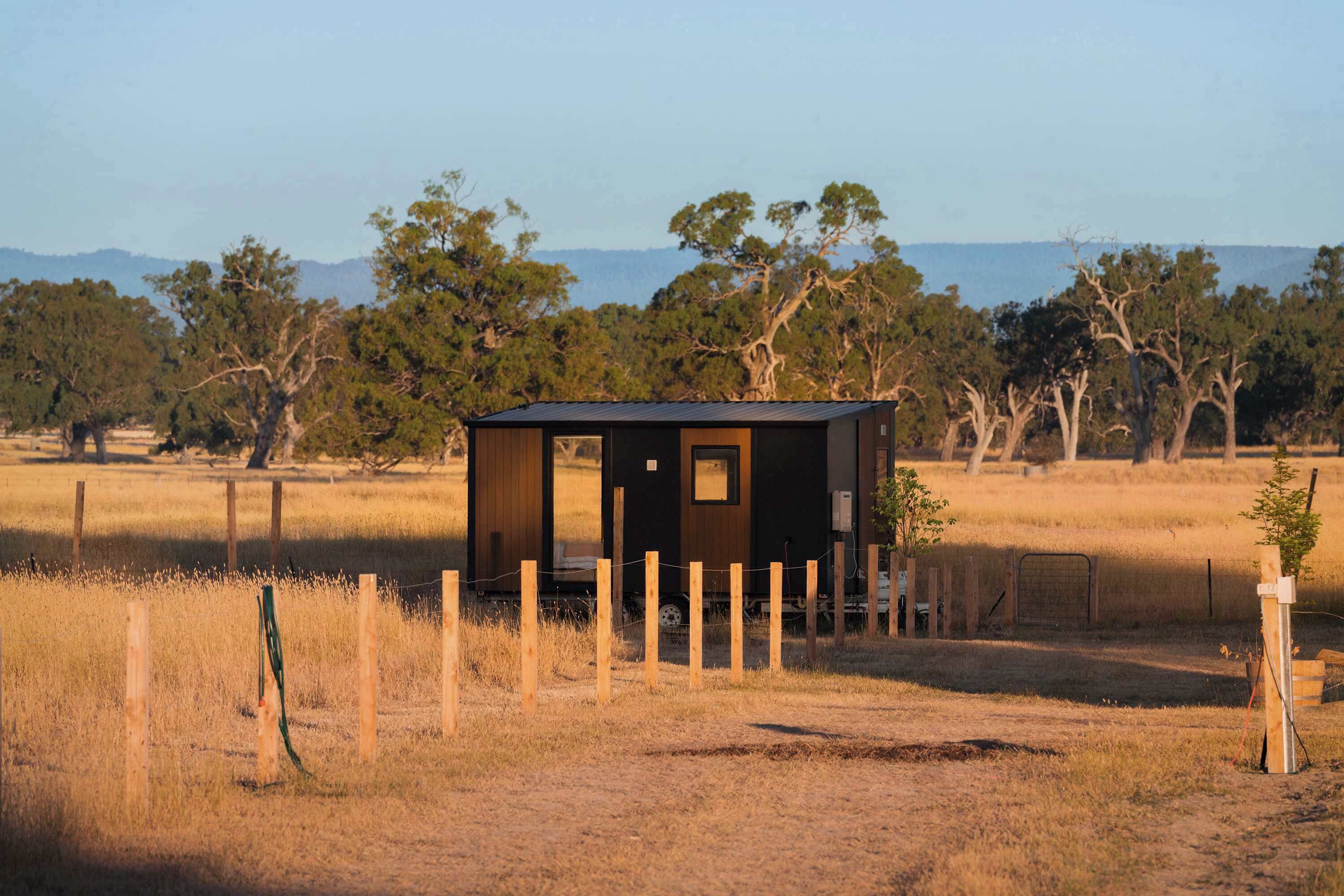 A small, single-story cabin with a dark exterior, surrounded by a wooden fence, set in a dry grass field with trees and mountains in the background.
