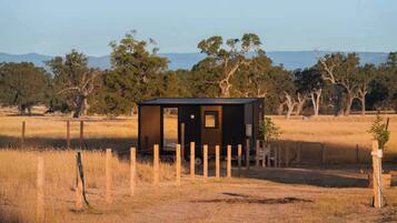 A small, single-story cabin with a dark exterior, surrounded by a wooden fence, set in a dry grass field with trees and mountains in the background.