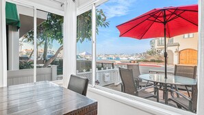 A balcony with a table and chairs, a red umbrella, and a view of a coastal area. - Bayfront 2BR with Stunning Harbor Views, Only Steps to the Beach! (Newport Beach)