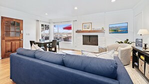 A living room with a blue sofa, a fireplace, a dining area with a table and chairs, and a view of the city through large windows. - Bayfront 2BR with Stunning Harbor Views, Only Steps to the Beach! (Newport Beach)