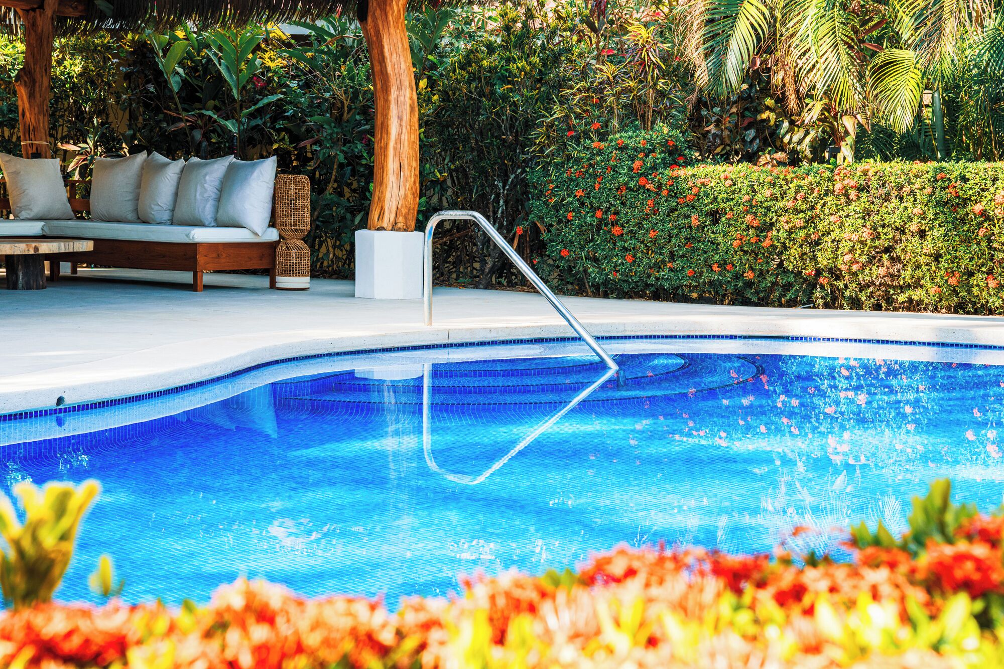 A swimming pool with a metal ladder, surrounded by lush greenery and a wooden deck with a sofa.