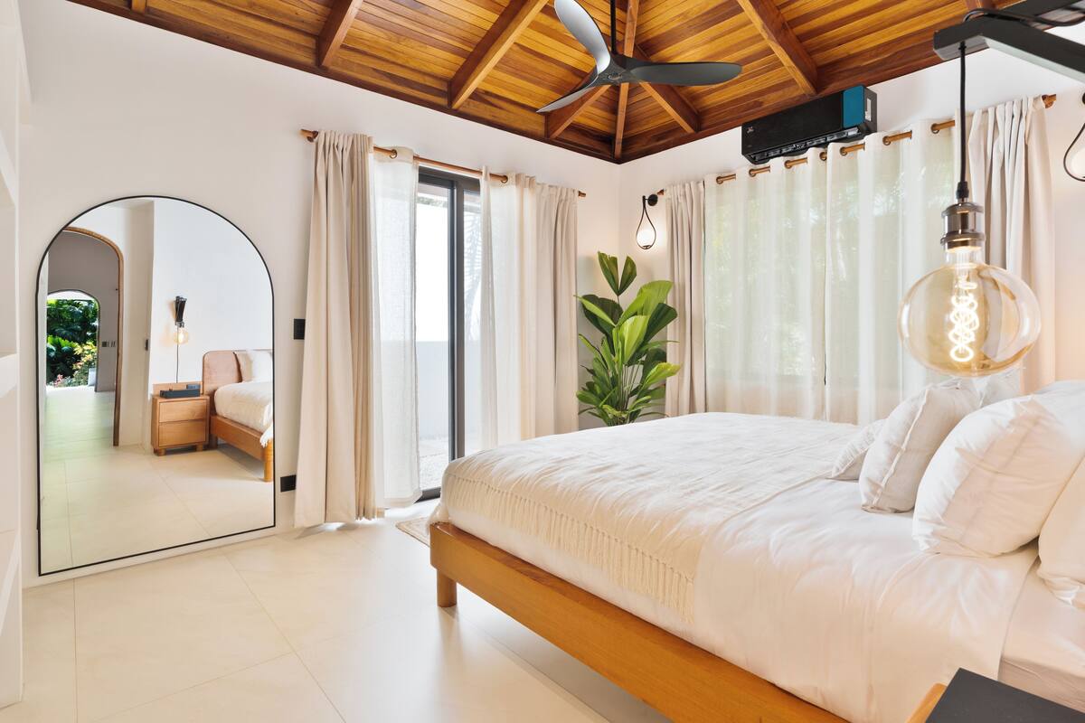 Une chambre avec un plafond en bois, un lit avec une literie blanche, un grand miroir et une plante en pot.