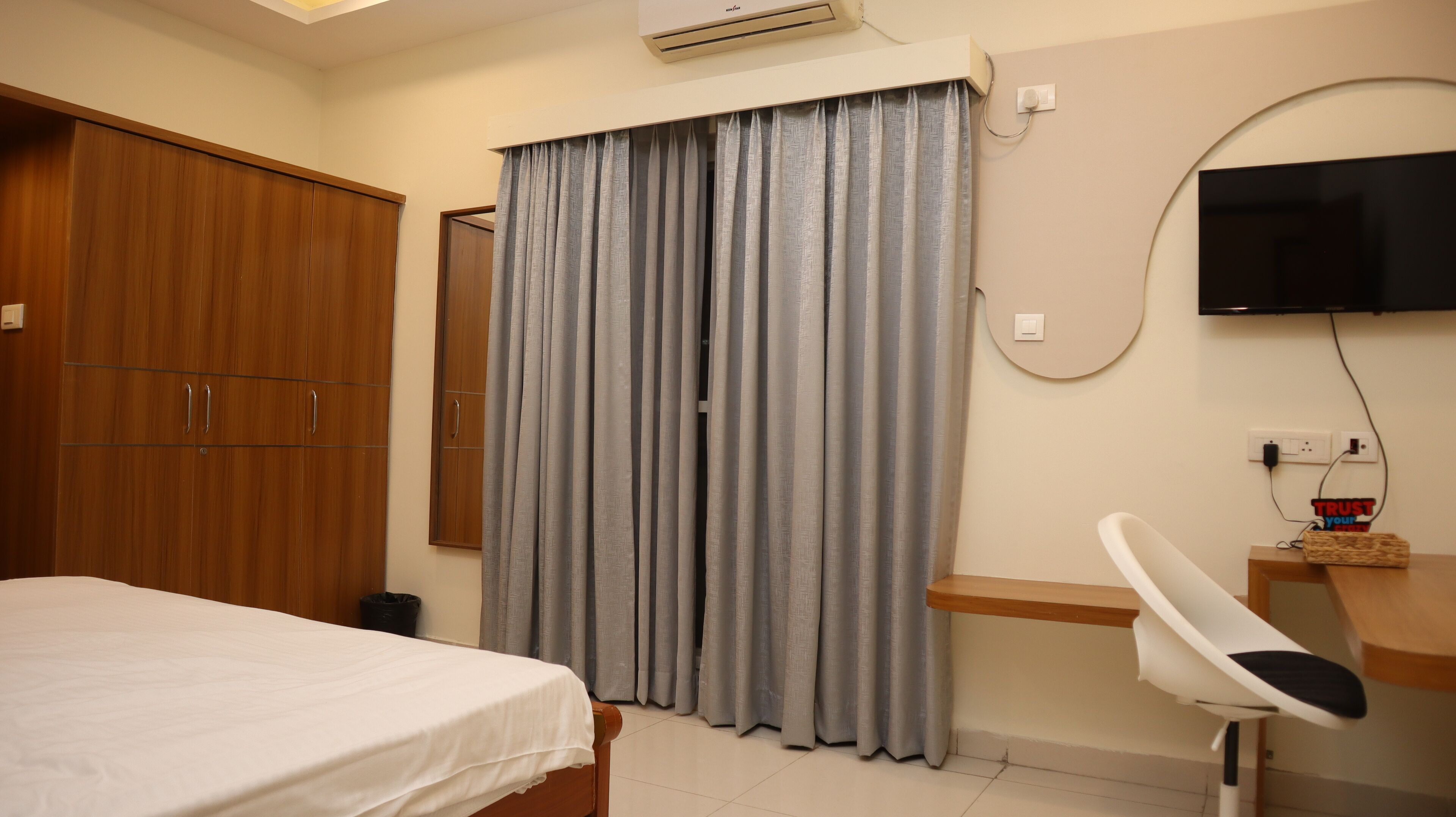 Une chambre d’hôtel comprenant un lit, une armoire en bois, un bureau avec une chaise, un téléviseur fixé au mur et des rideaux gris.