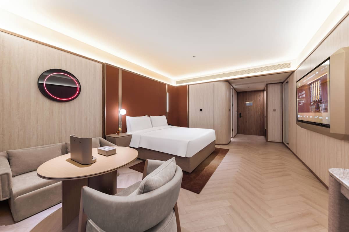 Een moderne hotelkamer met een bed, een zithoek met een tafel en stoelen, een aan de muur bevestigde televisie en een houten vloer.