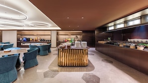 Una moderna hall d'albergo con un'area bar centrale, diverse disposizioni di posti a sedere e una cucina sullo sfondo.