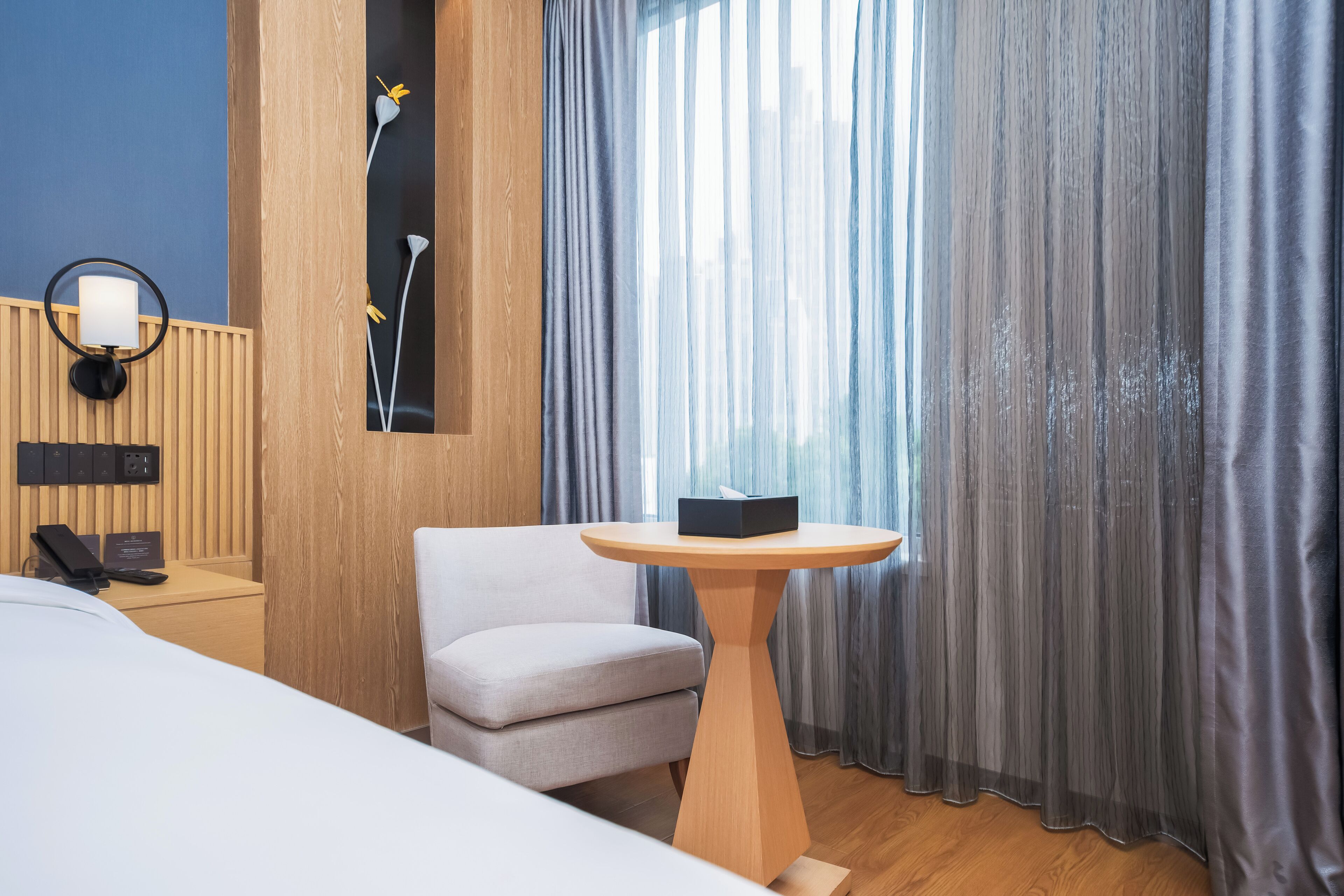 Et hotelværelse med en seng, et rundt bord med en kasse, en stol, en vægmonteret lampe og et vindue med lette gardiner.