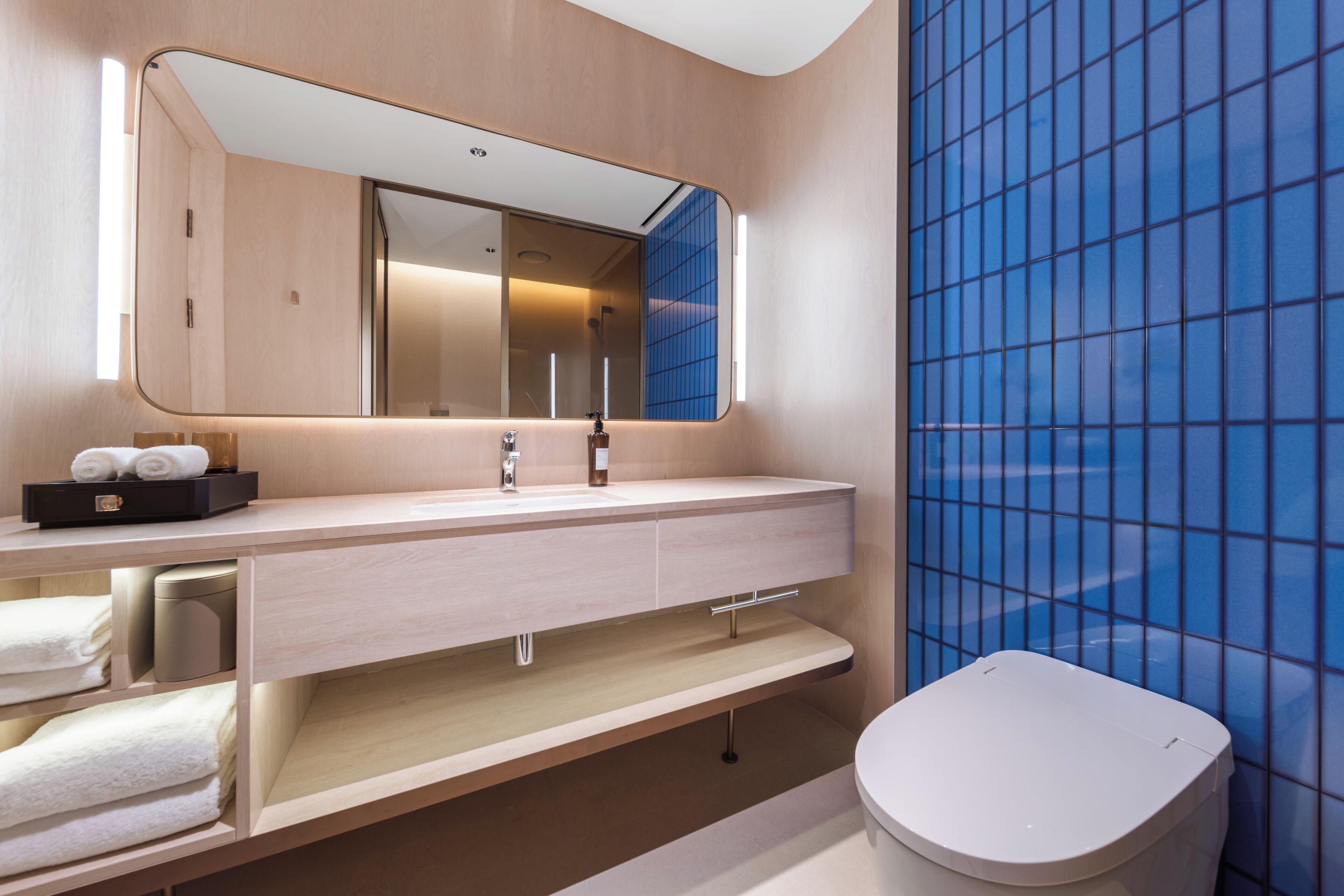 A modern bathroom with a large mirror, a sink, a toilet, and a shelf with towels.
