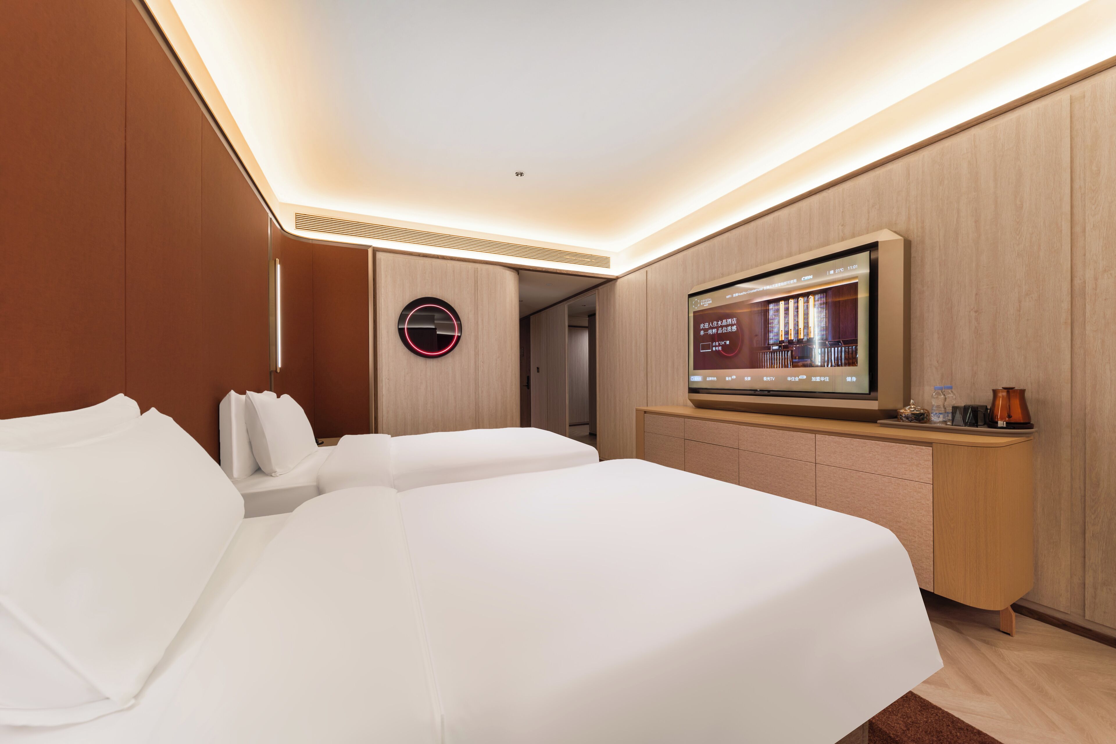 A modern hotel room with two beds, a flat-screen TV, a wooden cabinet, and a round clock on the wall.