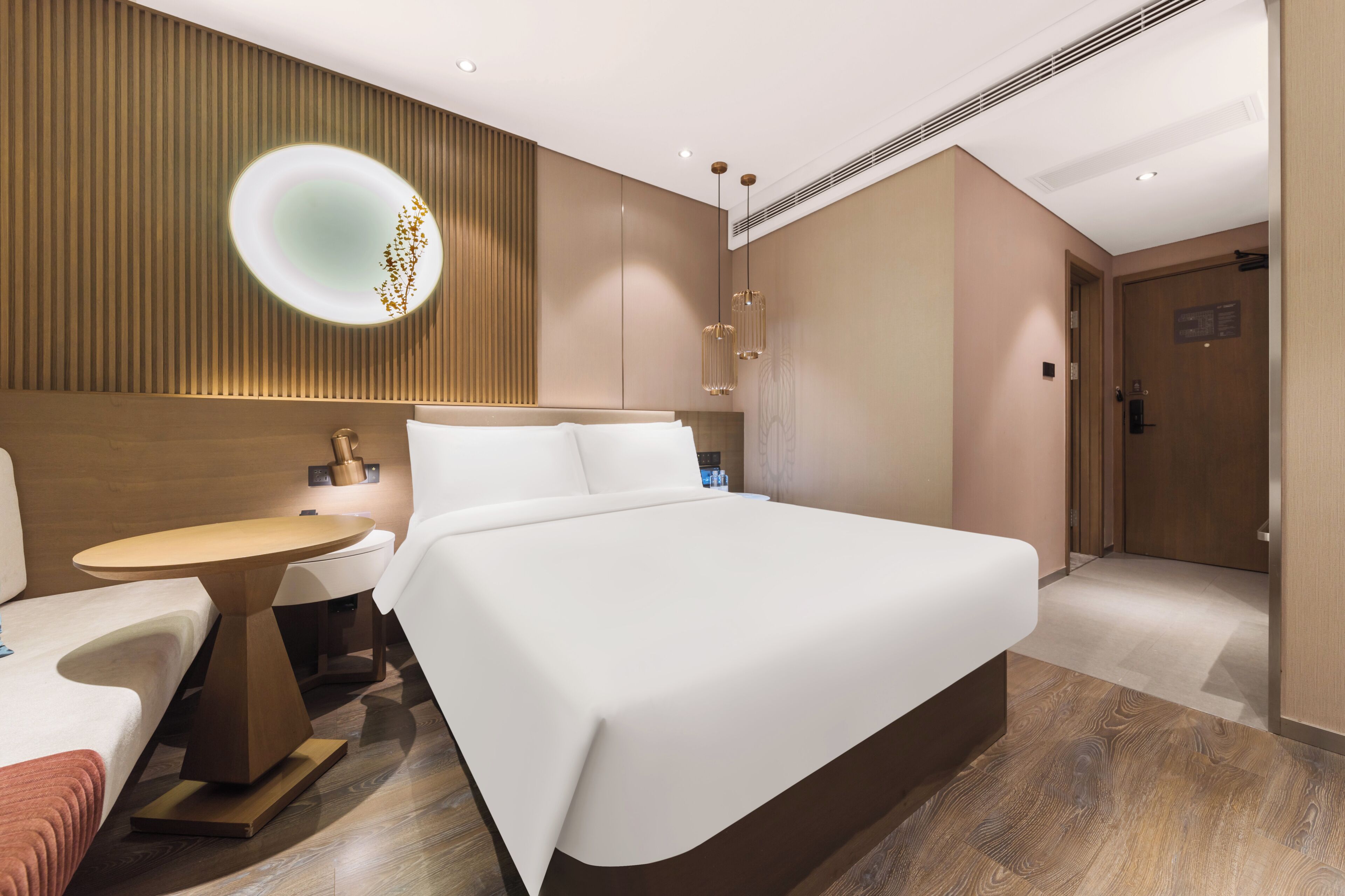 A modern hotel room with a large bed, bedside tables, a round table, and a wall-mounted light fixture.
