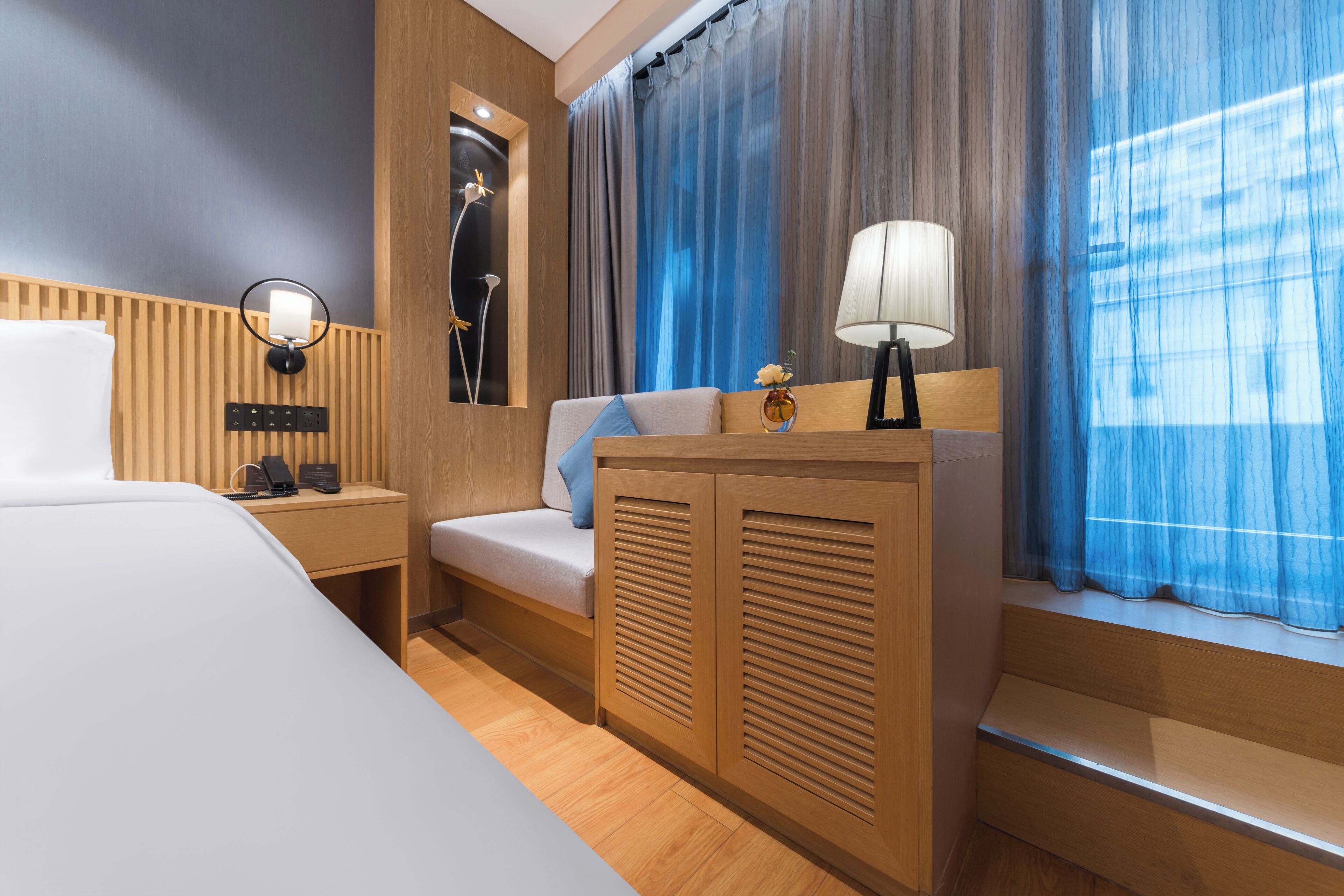 A modern hotel room with a bed, a desk with a lamp, a vanity with a mirror, and a window with curtains.
