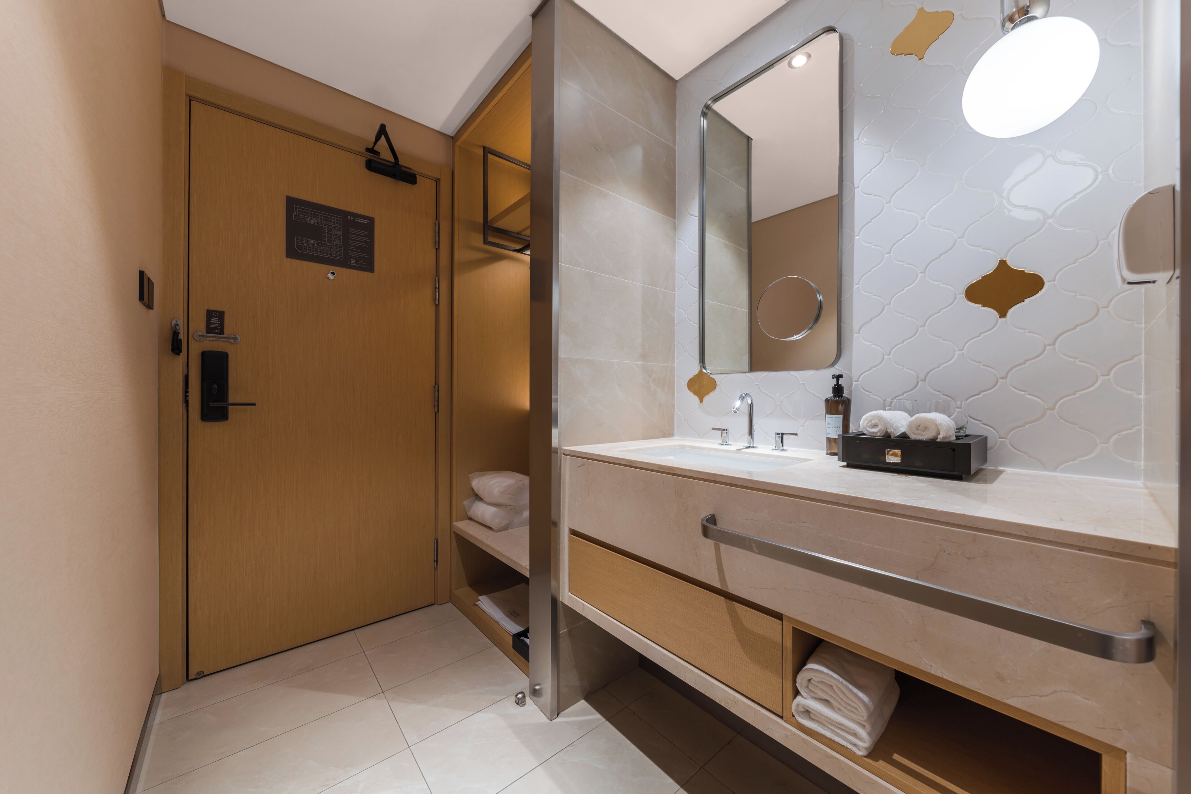 A modern bathroom with a wooden door, a large mirror, a sink with toiletries, and a towel rack.