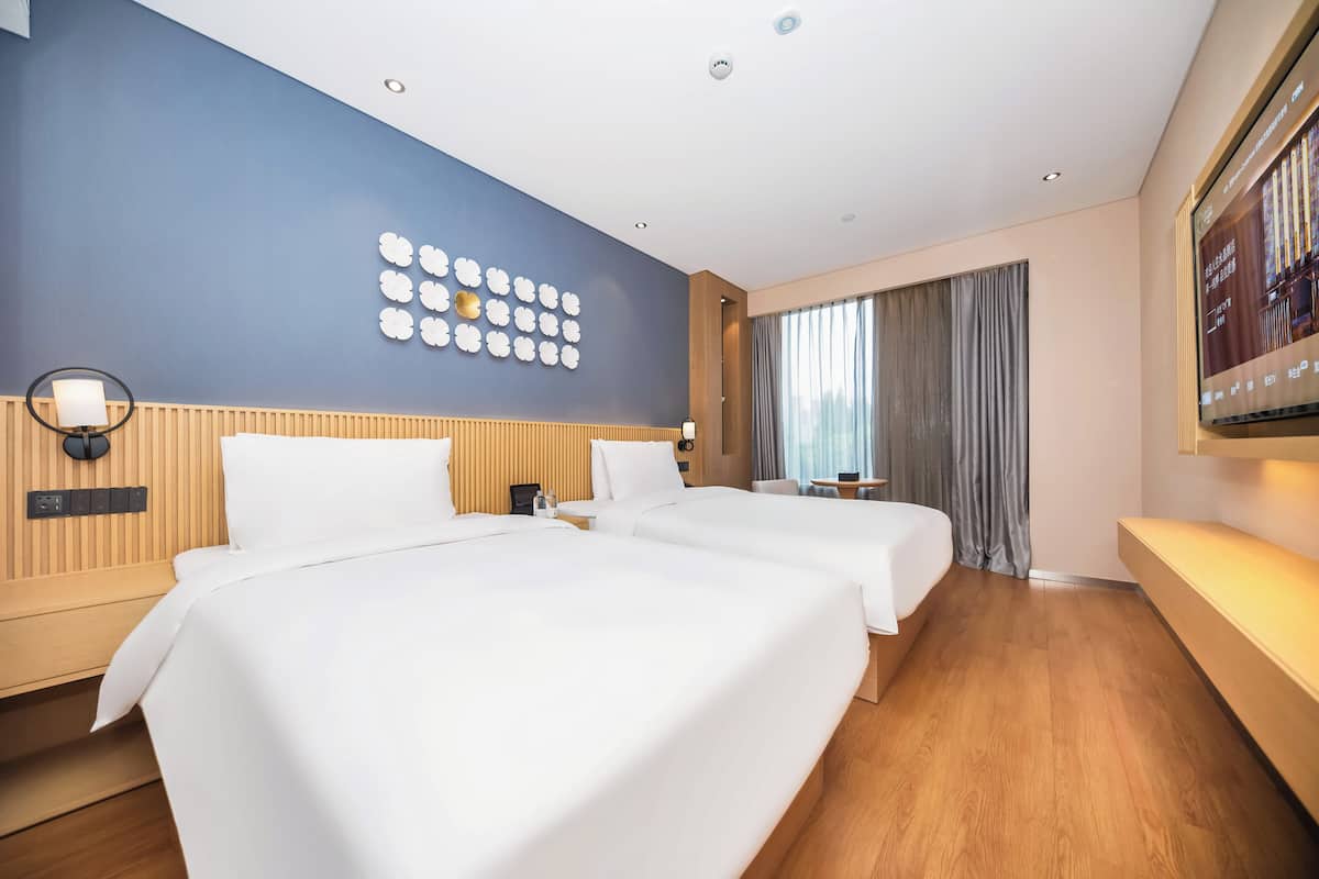 Een hotelkamer met twee bedden, een houten hoofdbord, een televisie aan de muur en een groot raam met gordijnen.
