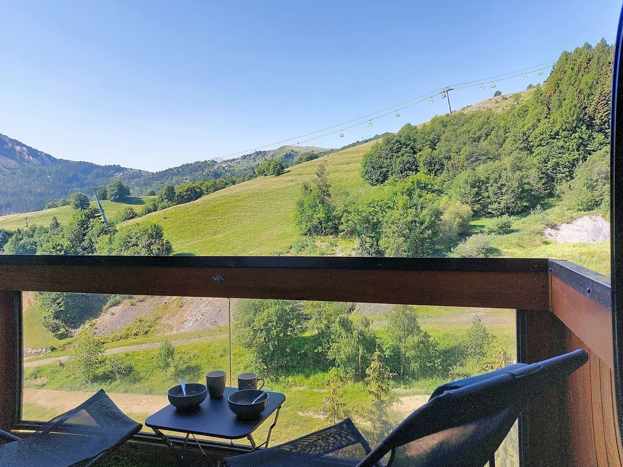 Un balcon avec une table et des chaises, donnant sur un paysage montagneux où l’on aperçoit, au loin, un téléphérique.