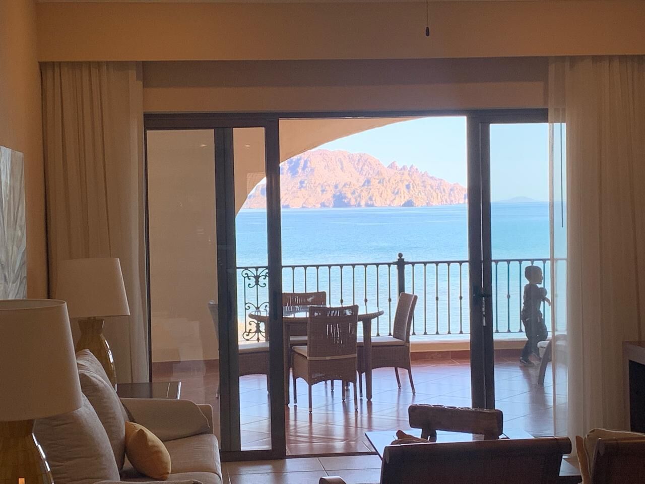 A room with a balcony overlooking a body of water and a mountain. There is a table with chairs and a sofa.