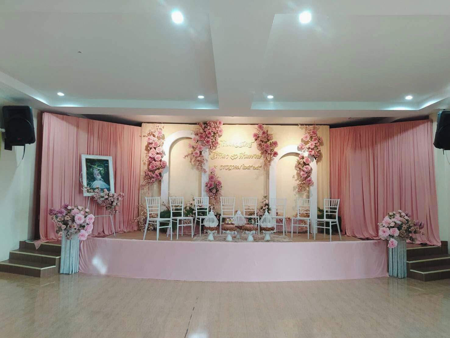 Un escenario con cortinas rosadas y decoraciones florales, sillas blancas y un cuadro en el lado izquierdo.
