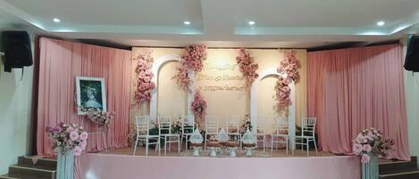 Un escenario con cortinas rosadas y decoraciones florales, sillas blancas y un cuadro en el lado izquierdo.