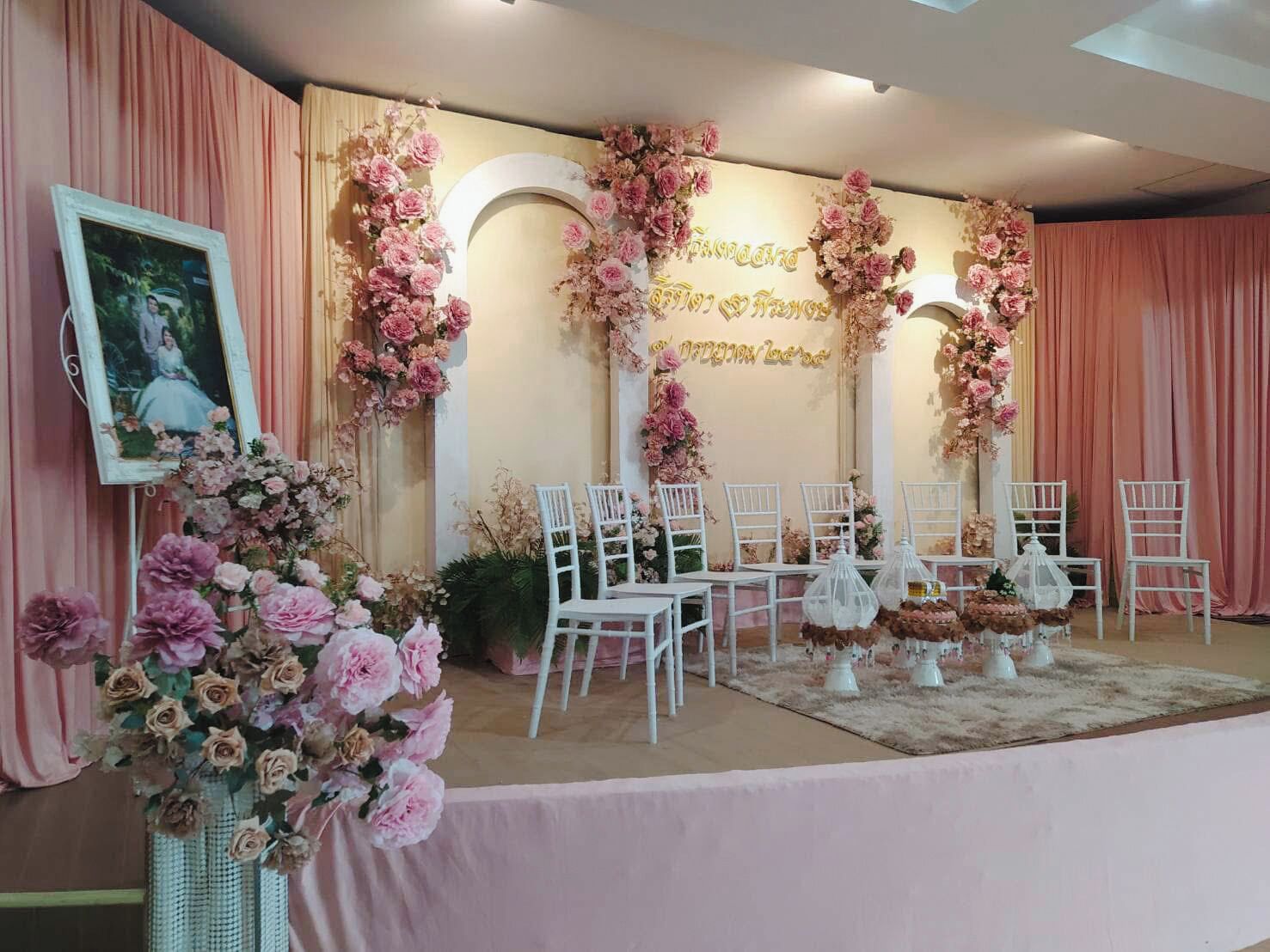 Un escenario con decoración floral, sillas blancas y un cuadro enmarcado.