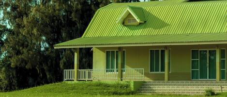 A single-story house with a green roof, a front porch with a railing, and a grassy yard.
