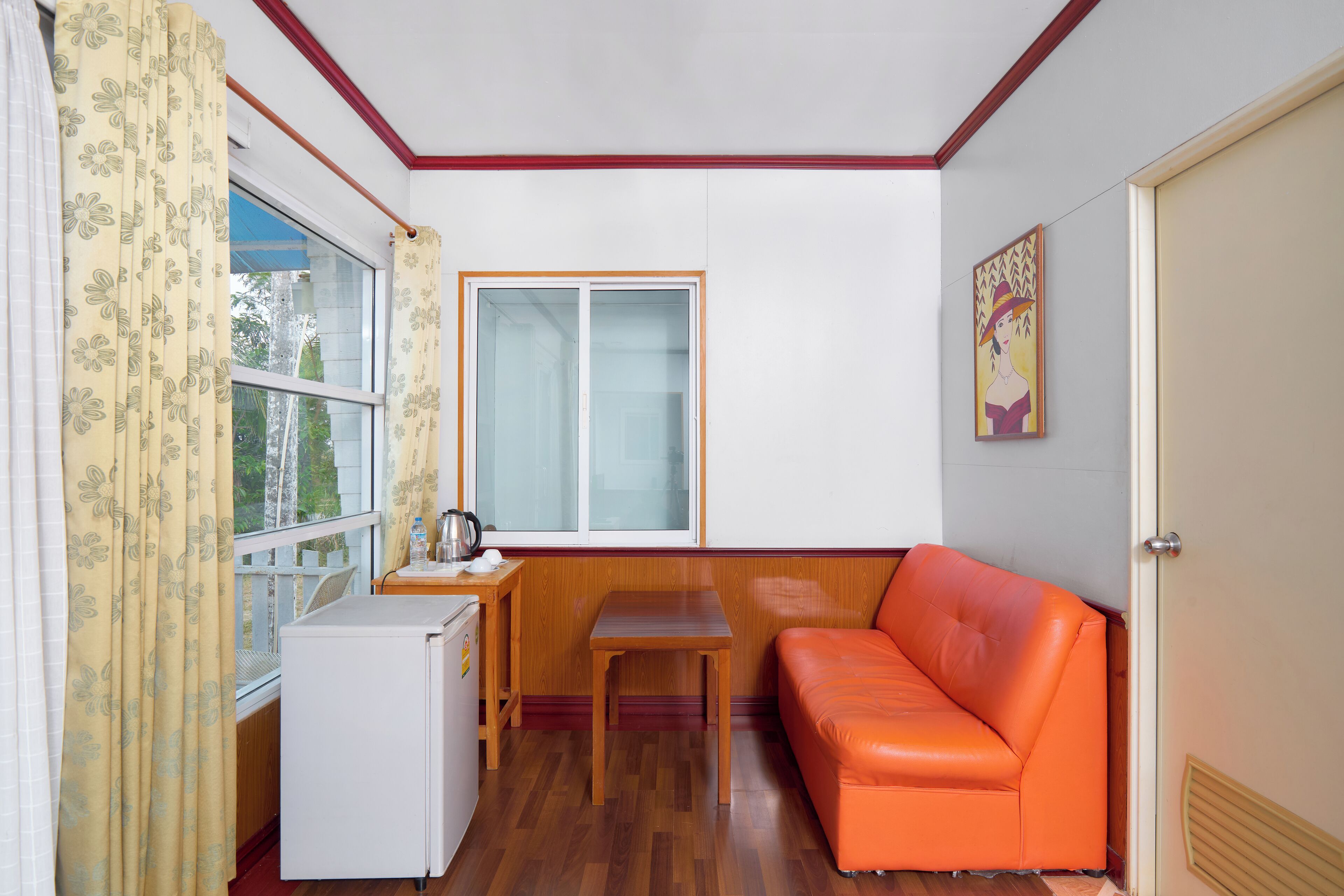 A compact room with a wooden table, a small white refrigerator, an orange sofa, and a window with curtains.