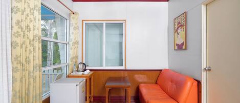 A compact room with a wooden table, a small white refrigerator, an orange sofa, and a window with curtains.