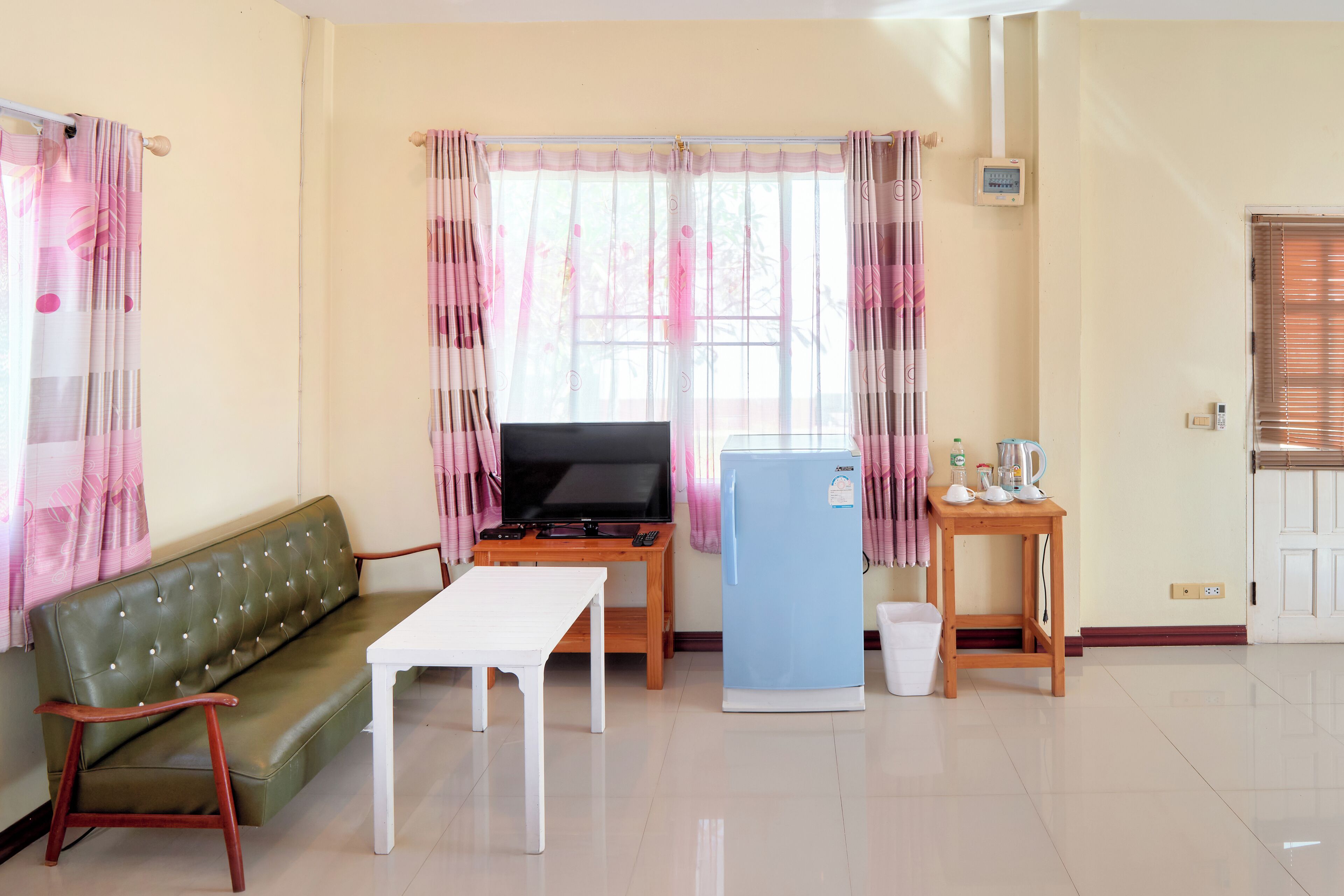 A living room with a sofa, a TV, a small table, a refrigerator, and a window with curtains.