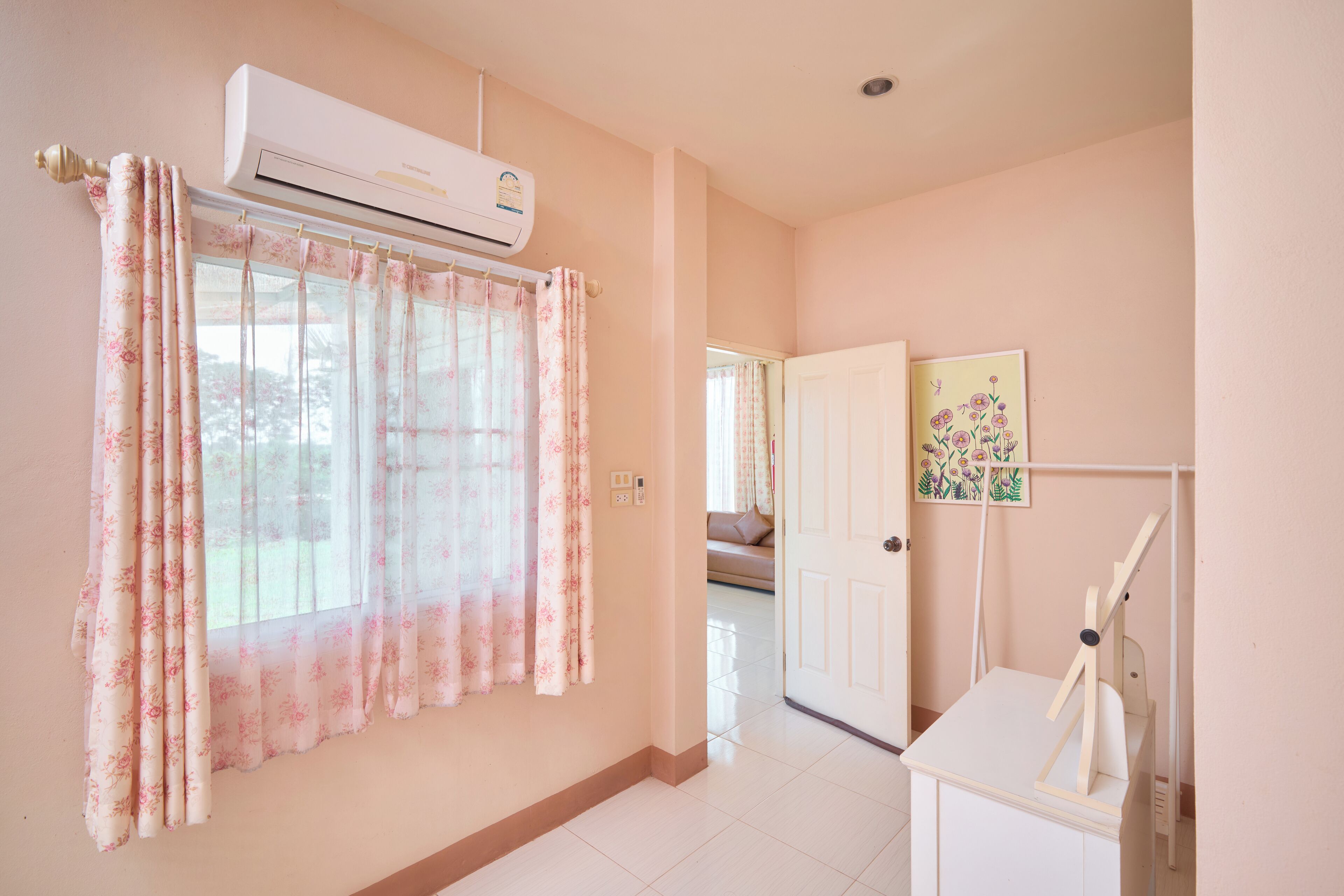 A room with a window covered by floral curtains, a white air conditioning unit, a white door, and a small table with a white chair.