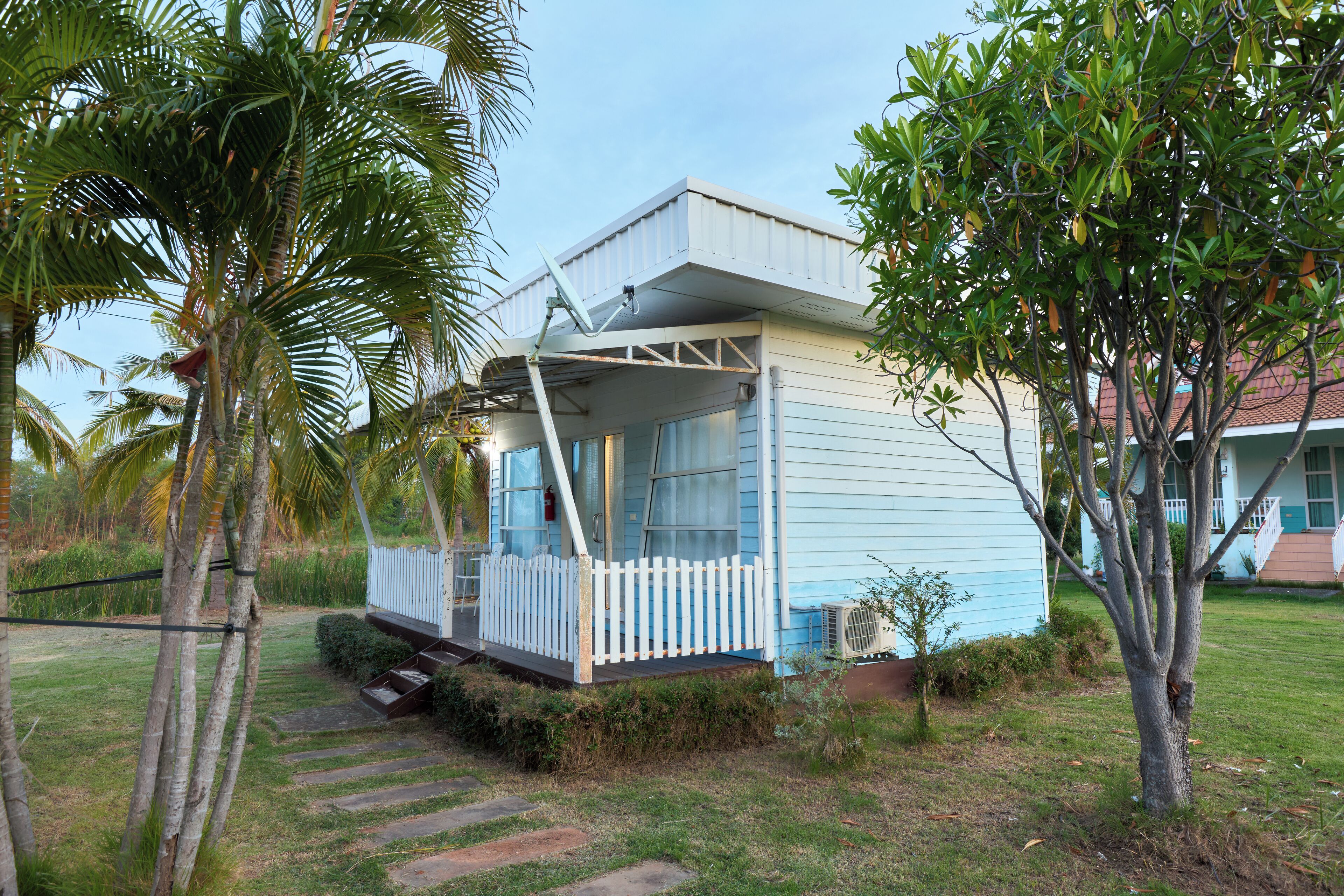 A small, single-story house with a porch and a small deck, surrounded by palm trees and a grassy yard.