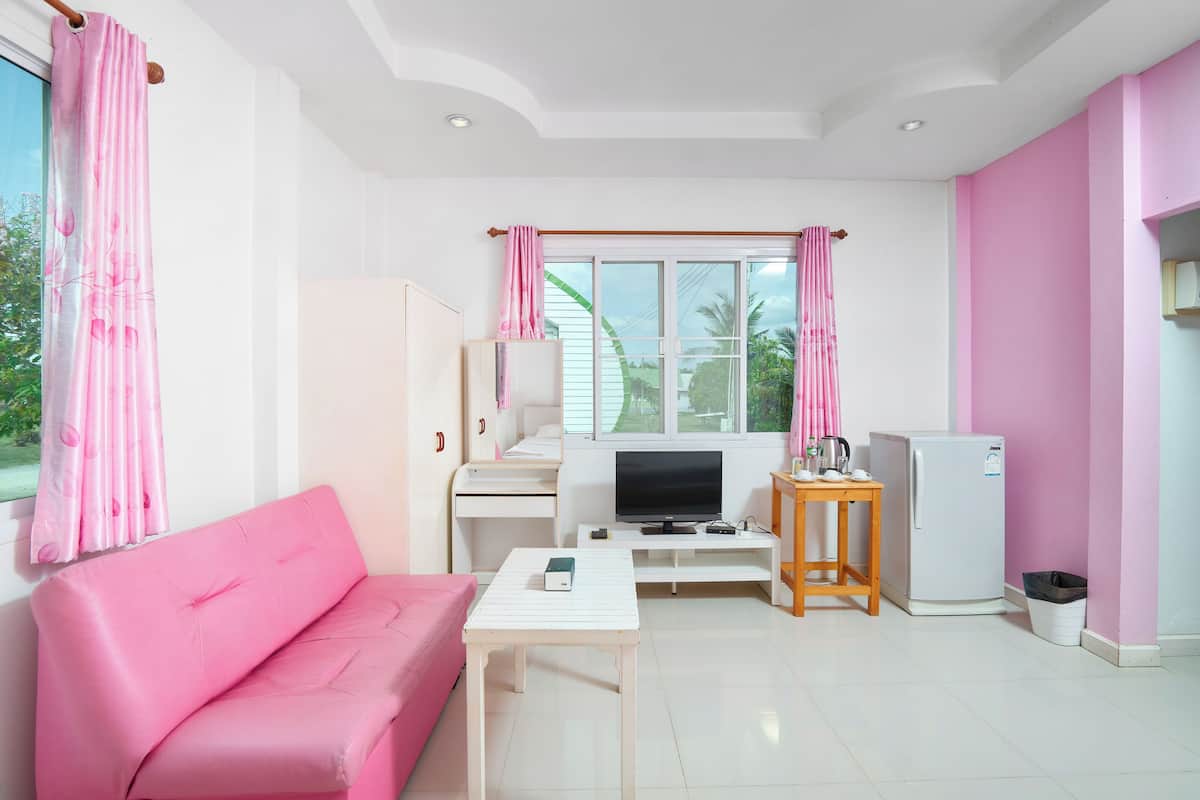 A room with a pink sofa, a white table, a TV, a refrigerator, and a window with curtains.