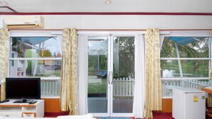 A room with a bed, a desk with a computer, a TV, and a refrigerator. There are curtains on the windows, and a view of a backyard with a fence and trees. - The Peak Resort Chaiyaphum (Chaiyaphum)