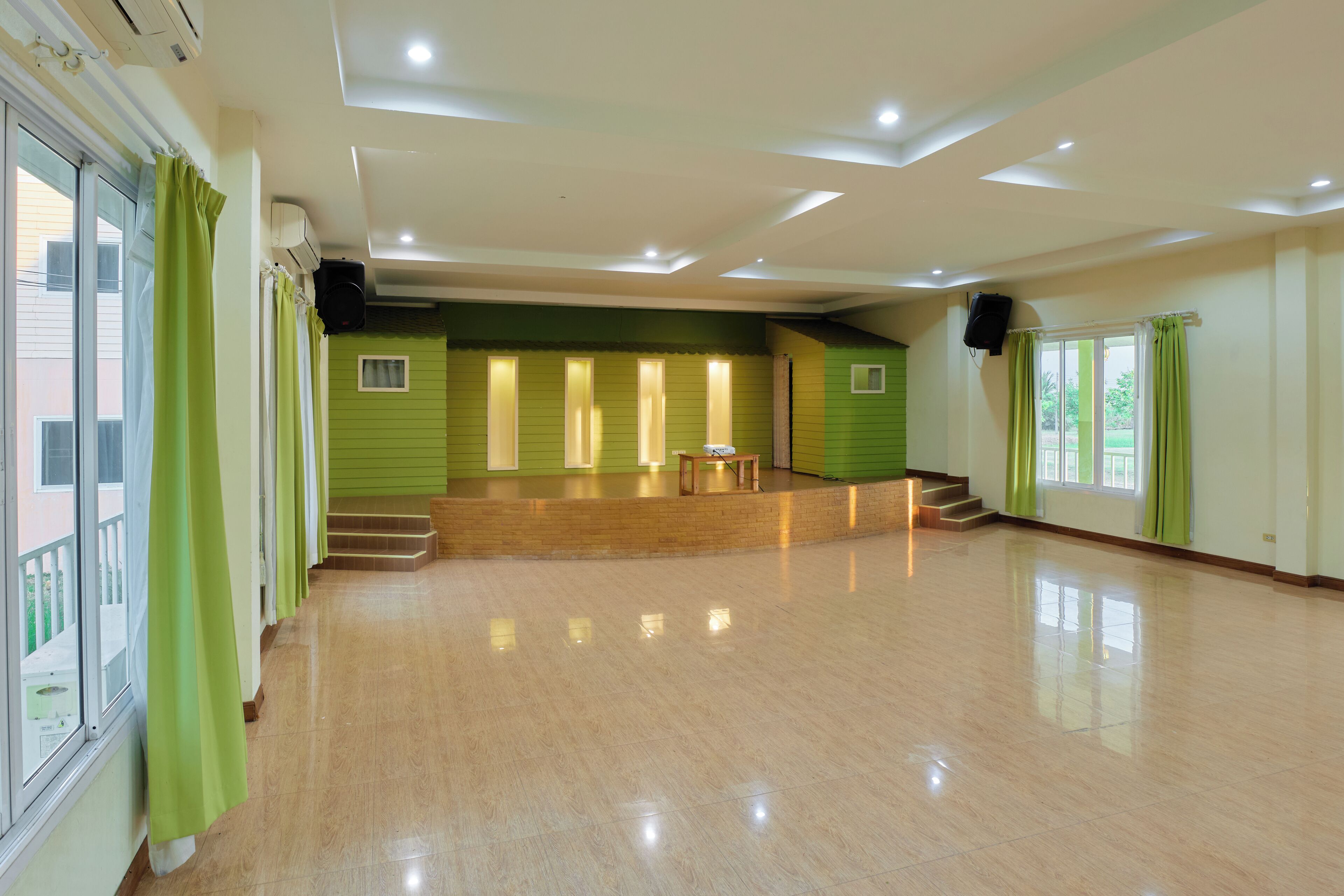 A spacious room with a polished wooden floor, a stage with a table, and a staircase leading to an upper level.
