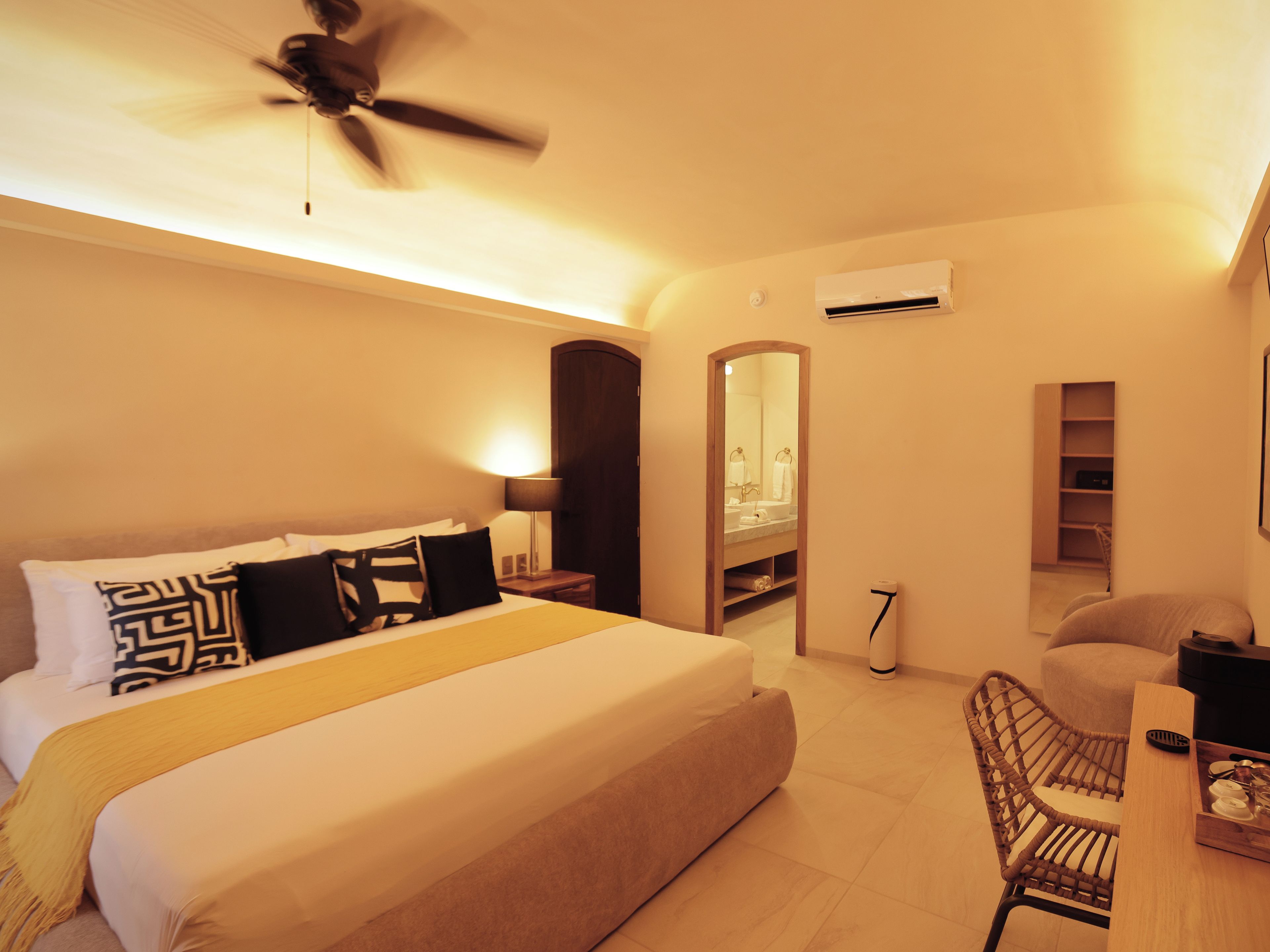 Habitación de hotel con cama, escritorio, silla, ventilador de techo, espejo y aire acondicionado.
