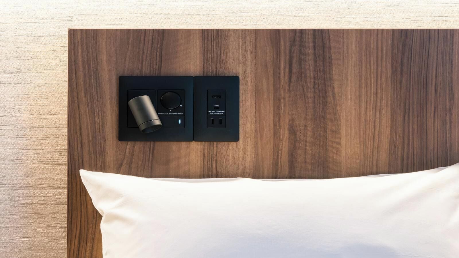A bed with a white pillow and a black bedside table with a cylindrical object and a USB port.