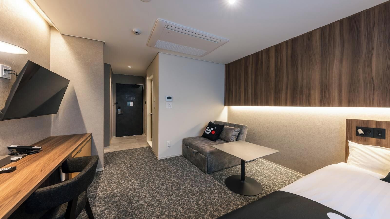 A hotel room with a bed, a desk with a chair, a small table, a sofa, and a TV mounted on the wall.