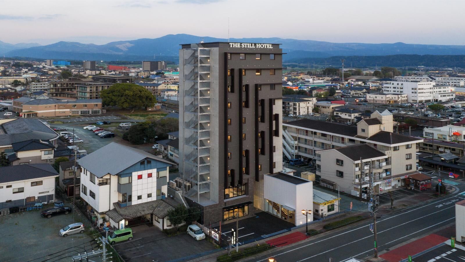A modern hotel with a central tower, surrounded by a parking lot and other buildings.