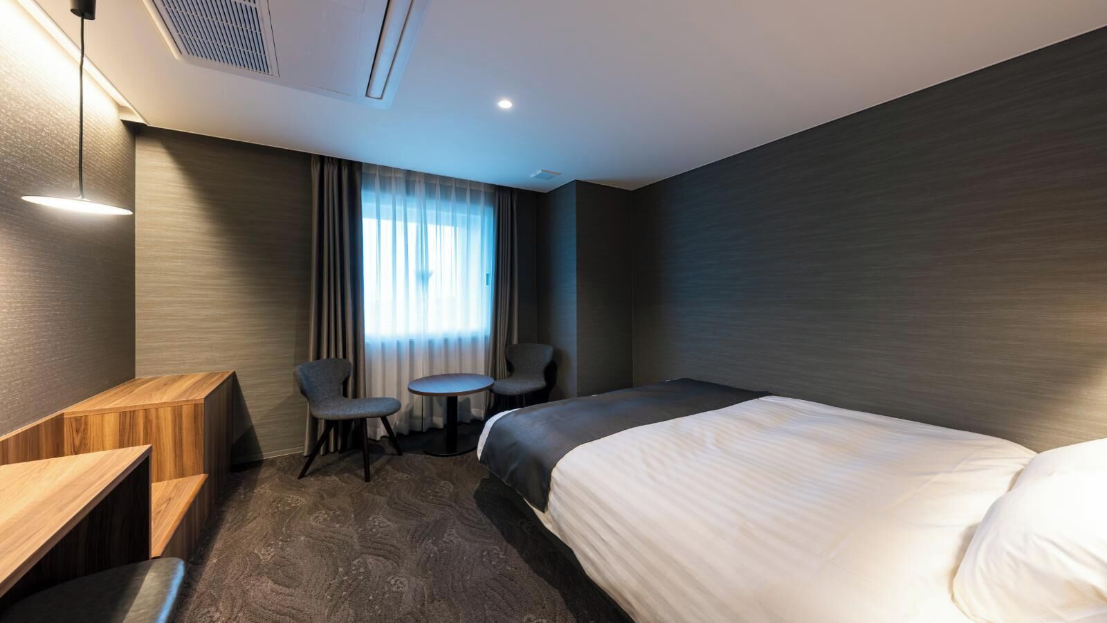 Quarto de hotel com uma cama, duas cadeiras, uma mesinha, uma escrivaninha e uma janela com cortinas.