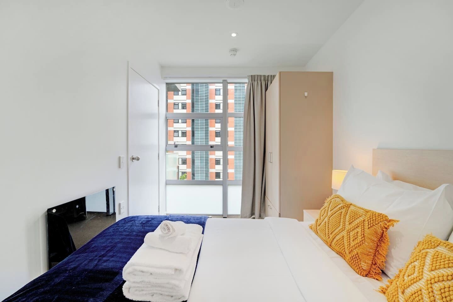 Une chambre d’hôtel avec un lit, une couverture bleue, un plaid jaune et une vue sur un immeuble par la fenêtre.