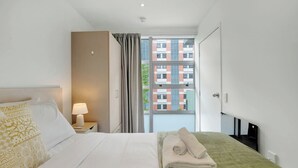 Hotelkamer met een bed, nachtkastje, lamp, raam met gordijnen en uitzicht op een gebouw.