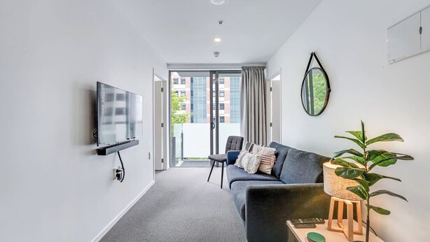 Een moderne woonkamer met een flatscreen televisie, een grijze bank, een klein houten tafeltje met een plant, en uitzicht op buiten door een glazen deur.