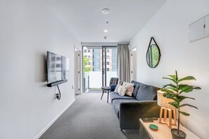Un salon moderne doté d’un téléviseur à écran plat, d’un canapé gris, d’une petite table en bois ornée d’une plante, et offrant une vue sur l’extérieur grâce à une porte vitrée.