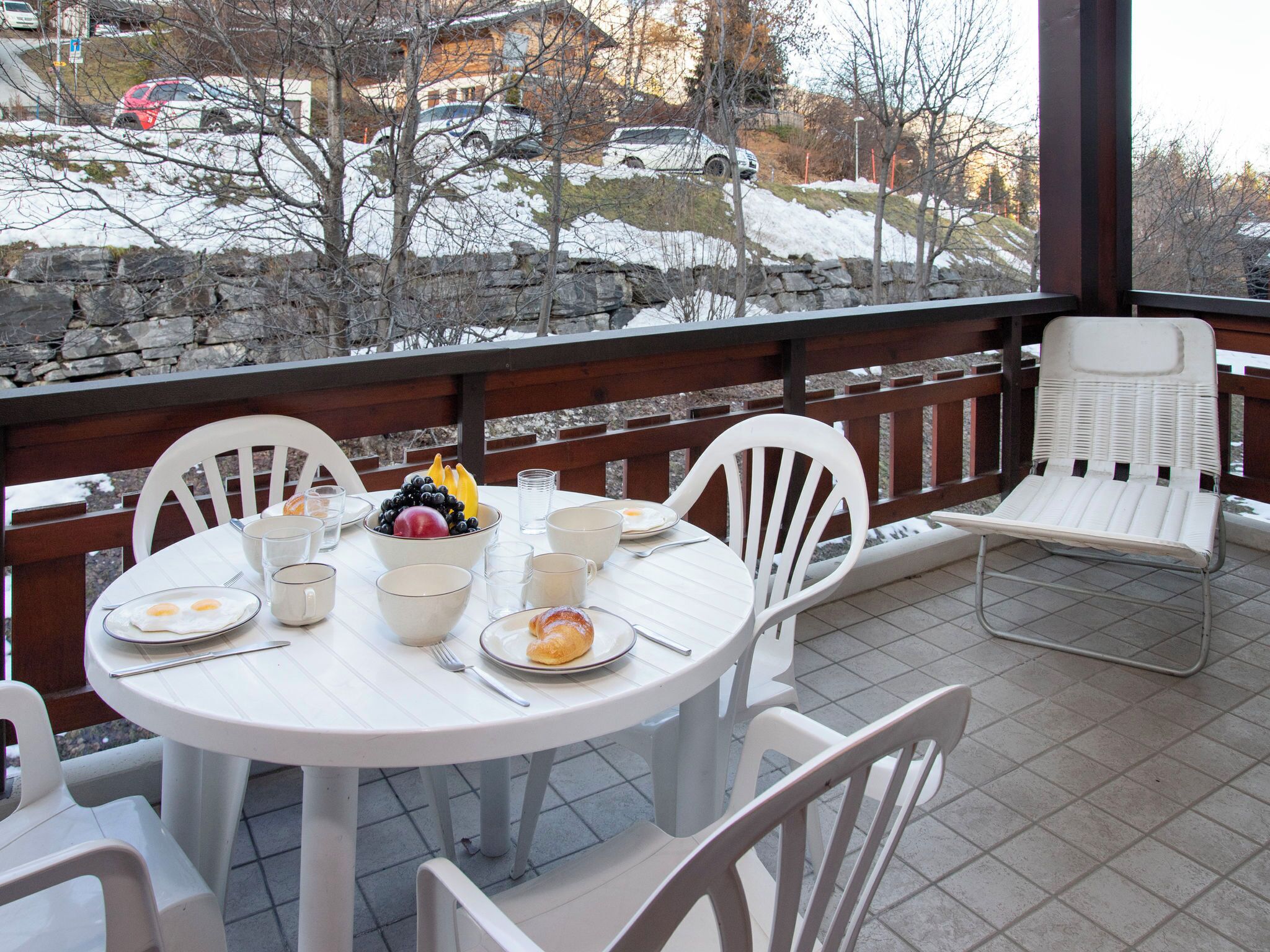 Un espace extérieur abrité, aménagé pour les repas, avec des tables et des chaises blanches, un mur en pierre et une vue sur un paysage enneigé où l’on aperçoit des voitures garées et des arbres.