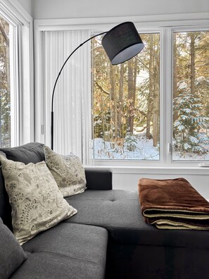 A modern living room with a grey sofa, patterned cushions, a brown throw blanket, and a black floor lamp. Large windows offer a view of a snowy forest. - Le Refuge du Mont | Cozy 1BR • Ski Getaway MSA (Beaupré)