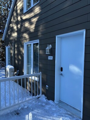 A house with a dark siding exterior, a white door, and a small porch with a white railing. - Le Refuge du Mont | Cozy 1BR • Ski Getaway MSA (Beaupré)