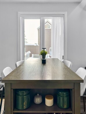 A dining area with a rectangular table, white chairs, and a view of a house through large windows. - Le Refuge du Mont | Cozy 1BR • Ski Getaway MSA (Beaupré)