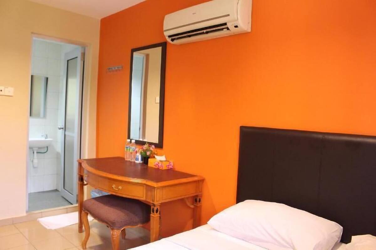A hotel room with an orange wall, a wooden desk with a mirror, a bed with a black headboard, and an air conditioning unit above the desk.
