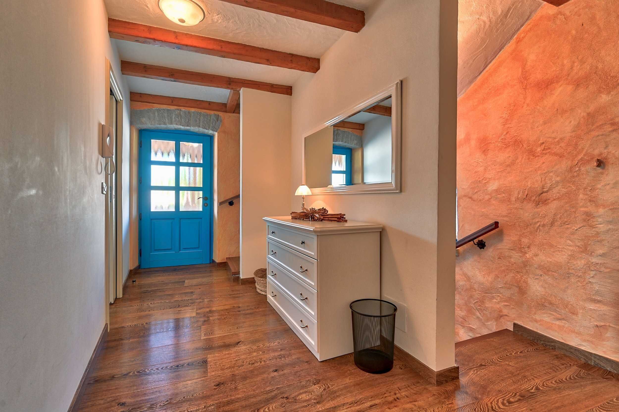 Un couloir avec un plancher en bois, une porte bleue, une commode blanche avec des tiroirs, un miroir et une poubelle.