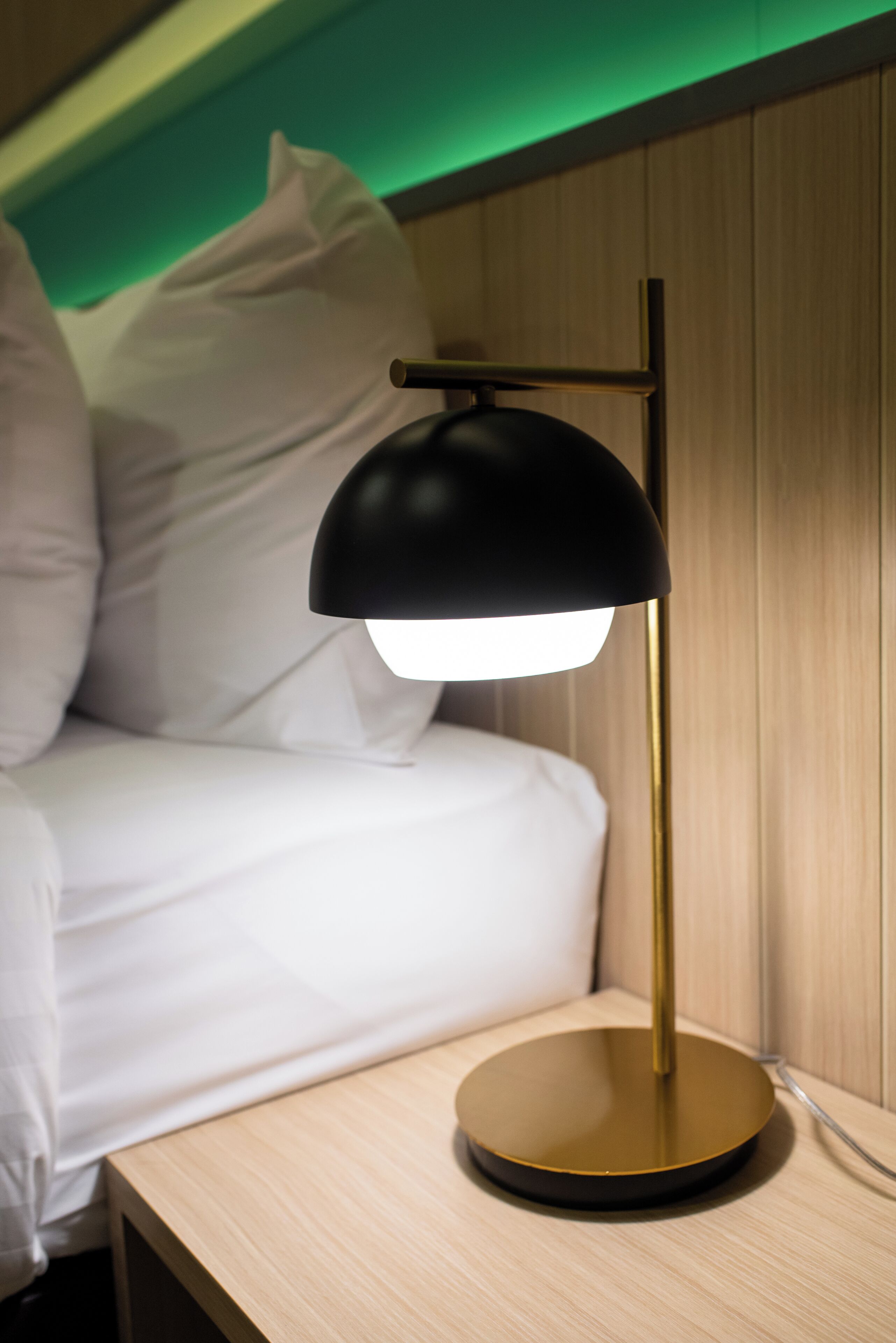 A bed with white bedding, a black and gold desk lamp, and a wooden nightstand.
