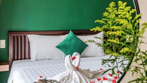Ein Bett mit weißen Leinen, einem grünen Kopfkissen und einem schwanenförmigen Handtuch. An der Wand über dem Bett hängen drei gerahmte Muster.