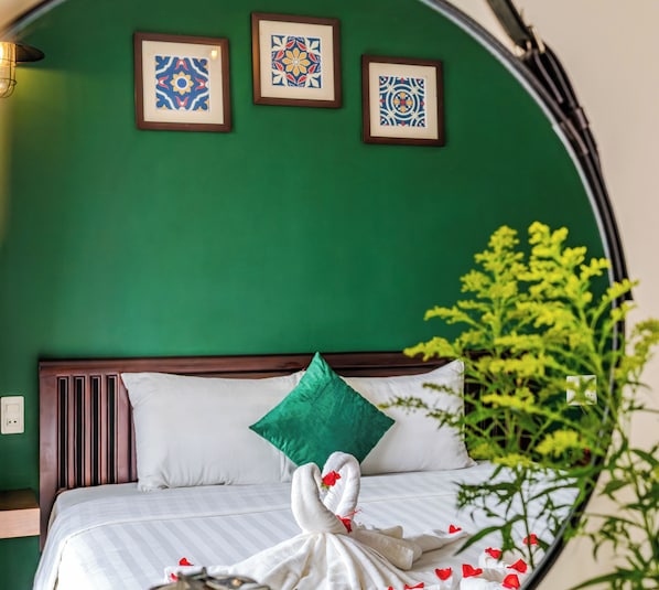 Ein Bett mit weißen Leinen, einem grünen Kopfkissen und einem schwanenförmigen Handtuch. An der Wand über dem Bett hängen drei gerahmte Muster.