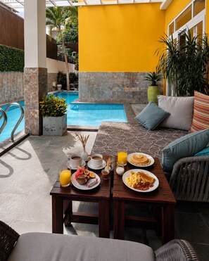 A poolside area with a dining table set for breakfast, a sofa with cushions, and a view of a swimming pool. - Horizon Hotel by LunaVerde Hoi An (Thanh pho Hoi An)