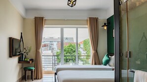 Ein Hotelzimmer mit Bett, Fernseher, einem kleinen Tisch mit einer Vase und Blick auf Gebäude und Bäume durch große Fenster.